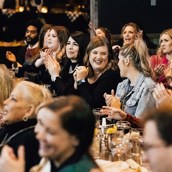 Audience at an event clapping and smiling, with a woman at center looking toward the stage.