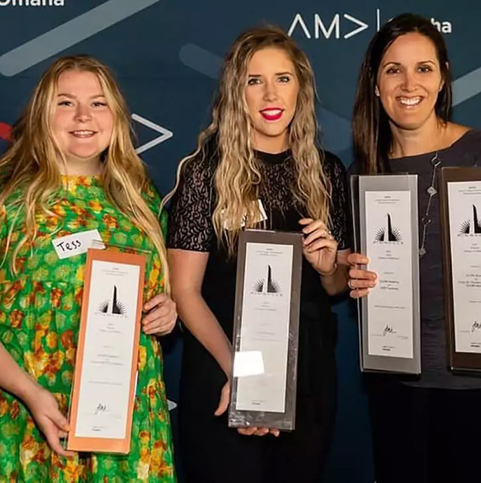 Three women standing in front of an AMA Omaha backdrop, each holding a tall award plaque.